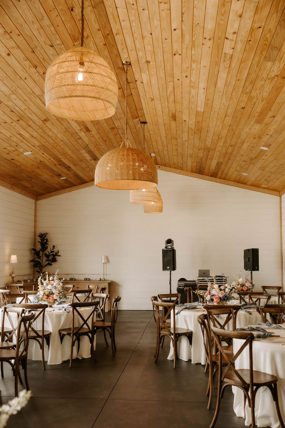 Reception hall with wooden ceiling, pendant lights and cross-back chairs set for a wedding.
