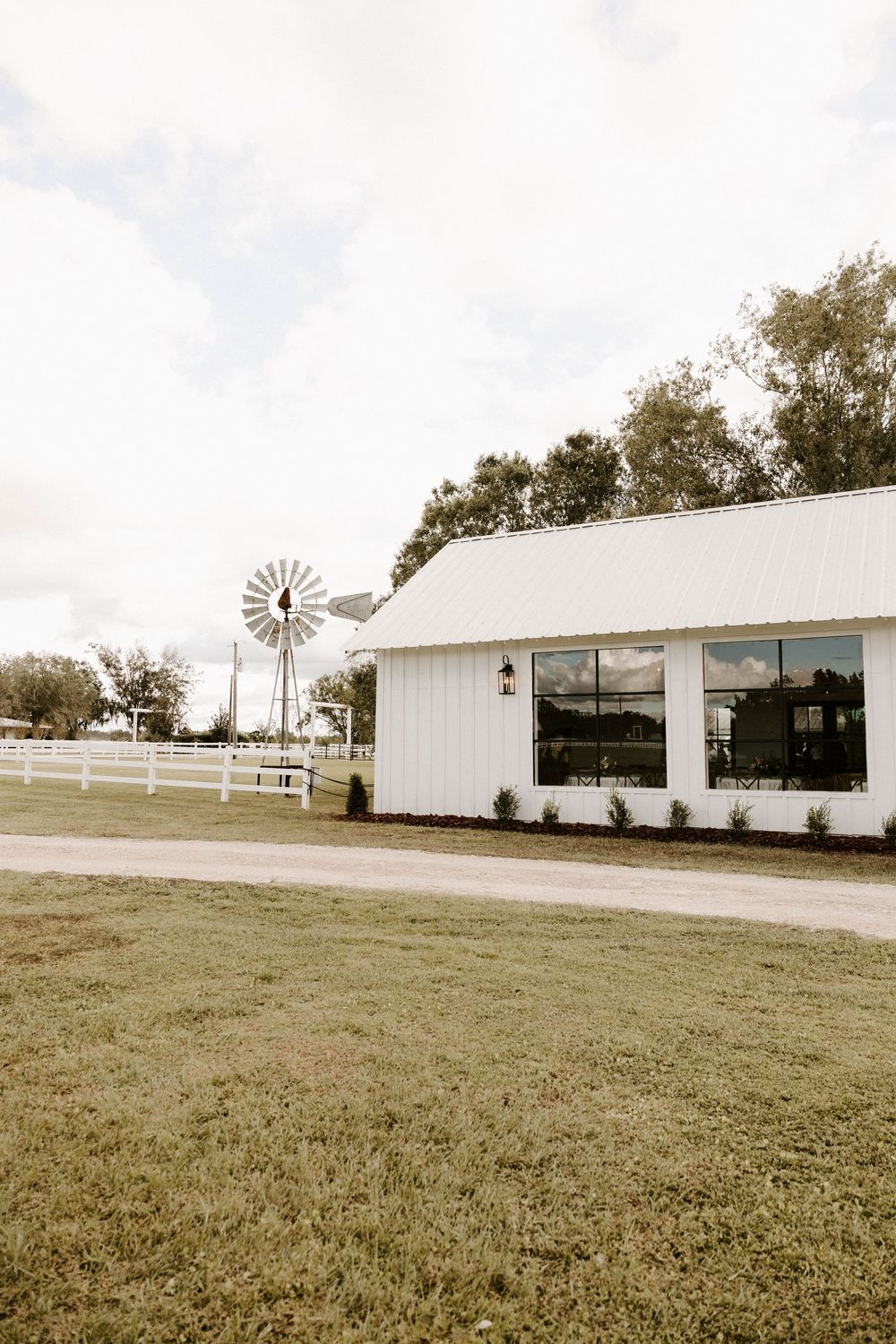 Panoramic view of a white modern farmhouse wedding venue with windmill and fenced grounds.