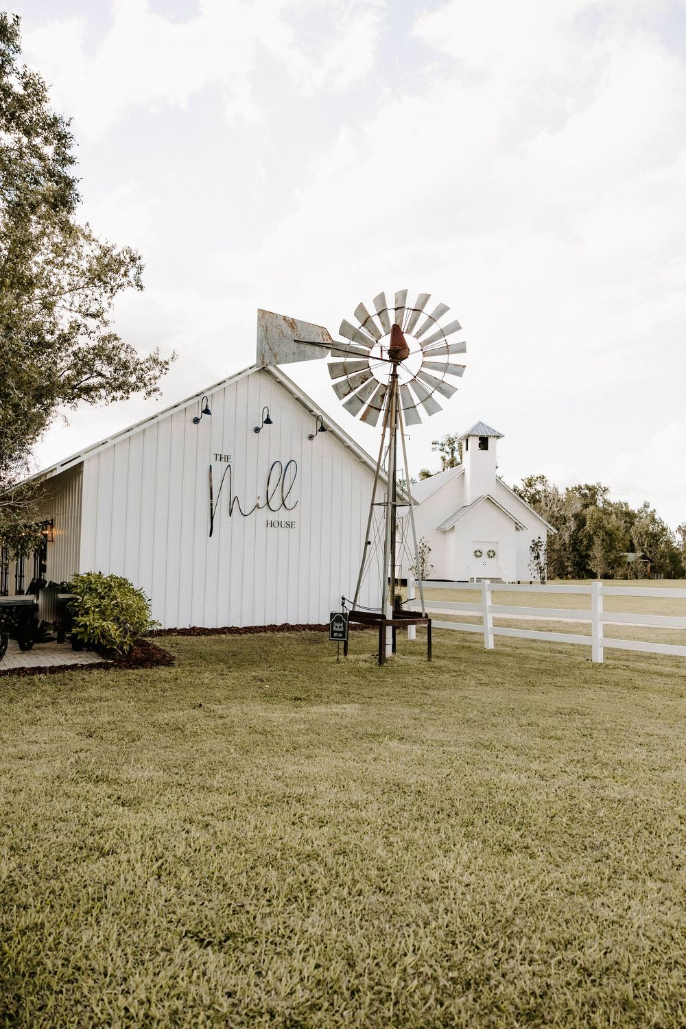Rustic white barn venue with vintage windmill and white fencing on a grassy hill.