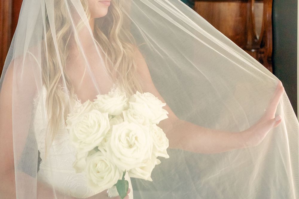Bride in lace gown holds a delicate white veil against a wooden interior.