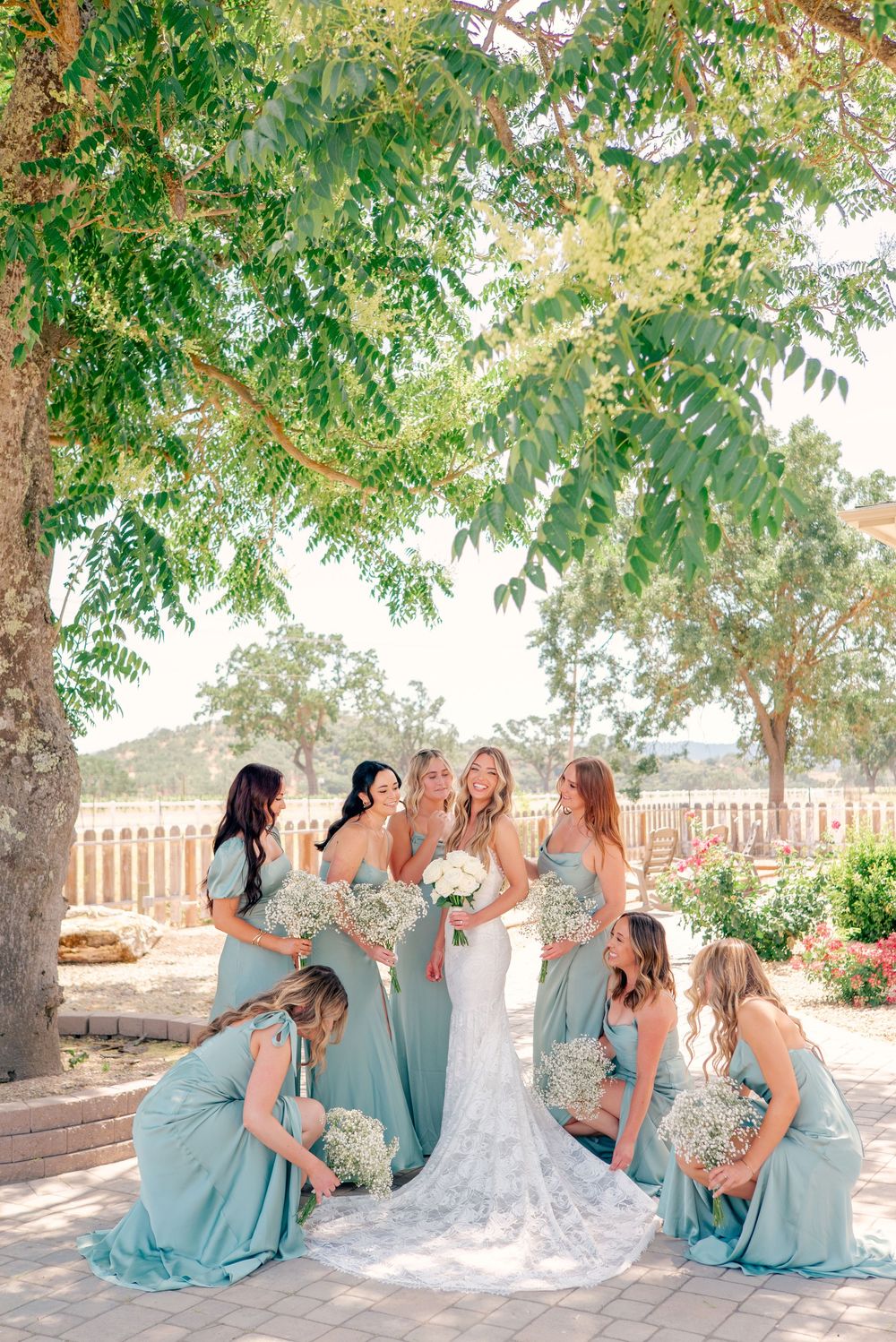 Wedding party in light blue bridesmaid dresses sits together outdoors under green trees.