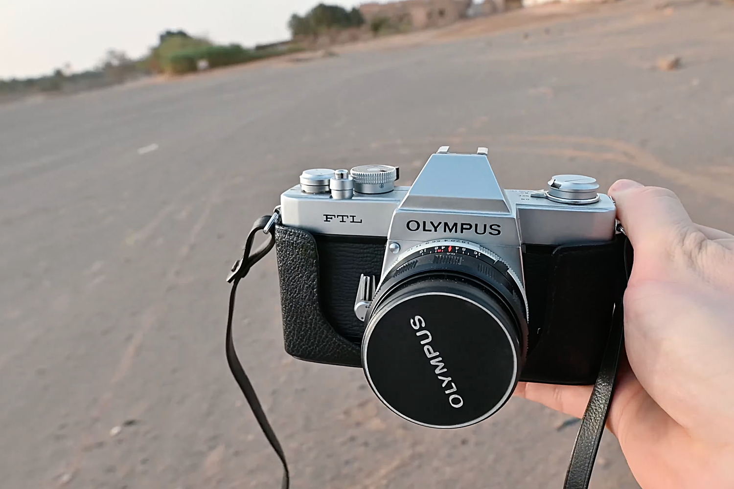 A hand holds a vintage Olympus film camera with lens against a blurred asphalt road background.