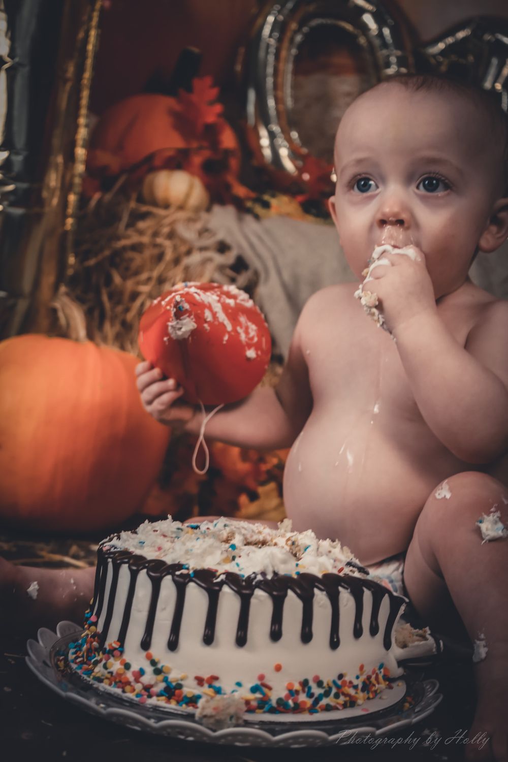 A cake smash photo session featuring a decorated white birthday cake with sprinkles and pumpkins in the background.