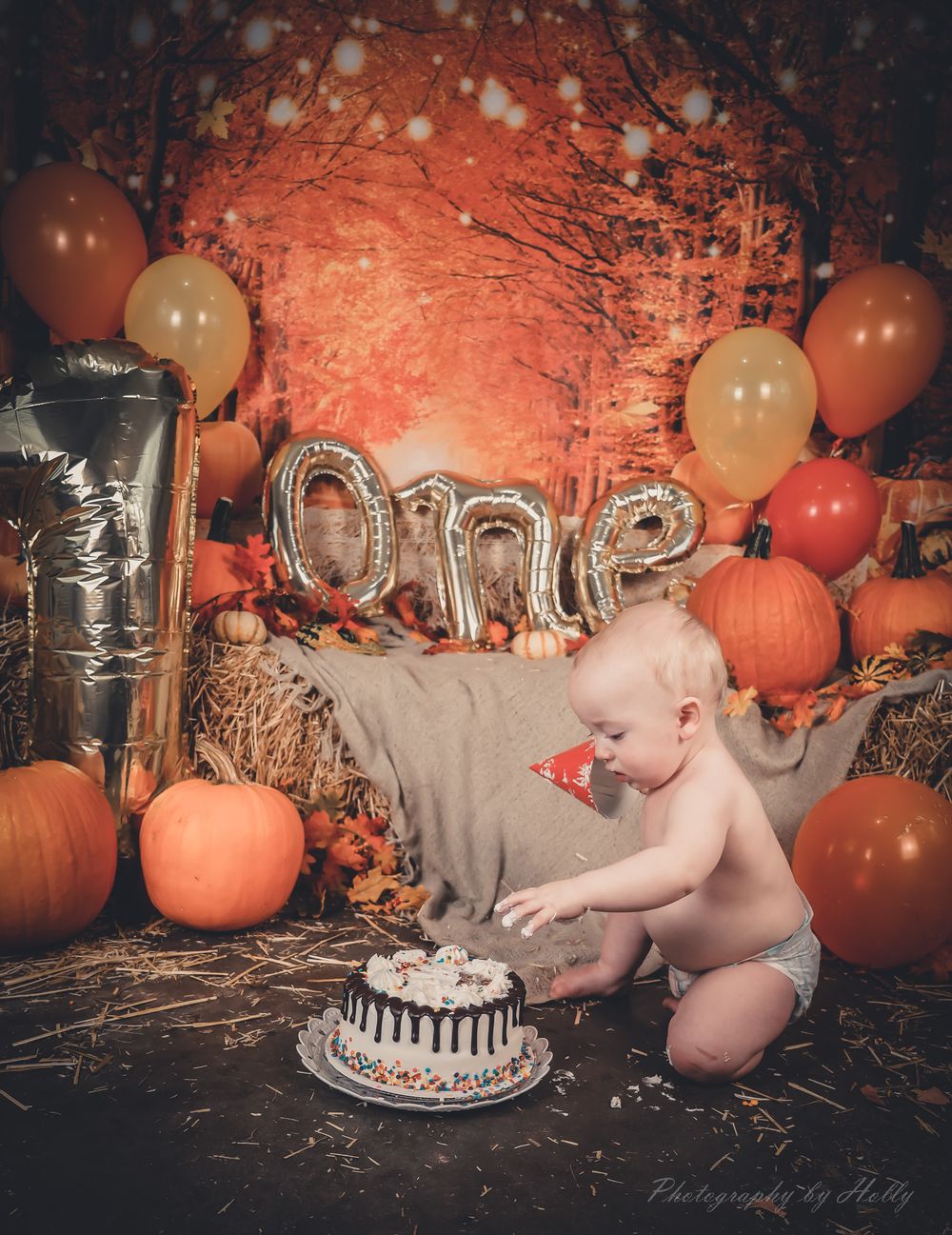A festive fall cake smash photo session with pumpkins, hay bales, and decorative number balloons in warm autumn lighting.