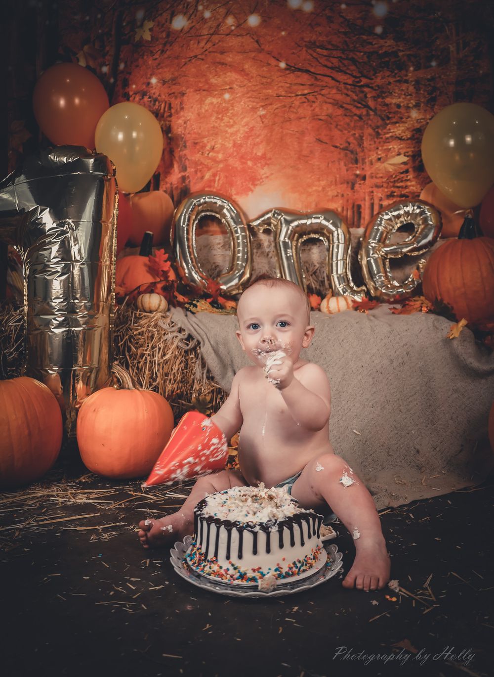 Festive autumn cake smash photo session with pumpkins, balloons, and silver letter decorations in rustic setting.