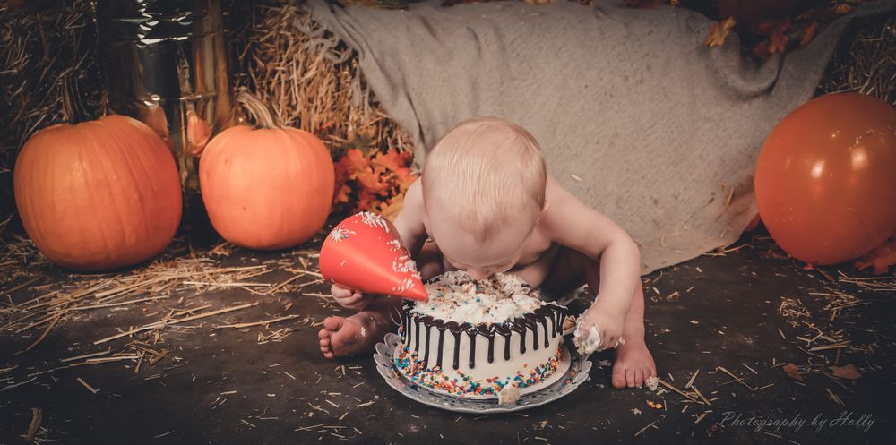 Baby smashing cake surrounded by pumpkins and fall decor in rustic autumn setting.