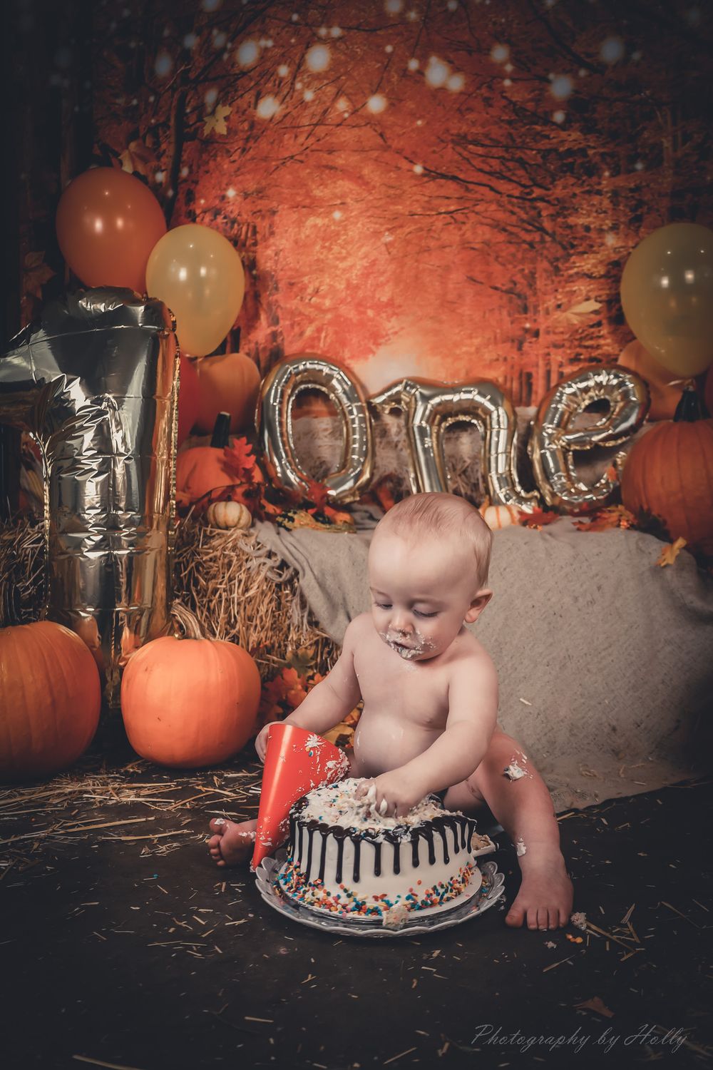 Baby enjoying cake smash photoshoot with pumpkins and silver balloons in dark moody setting.