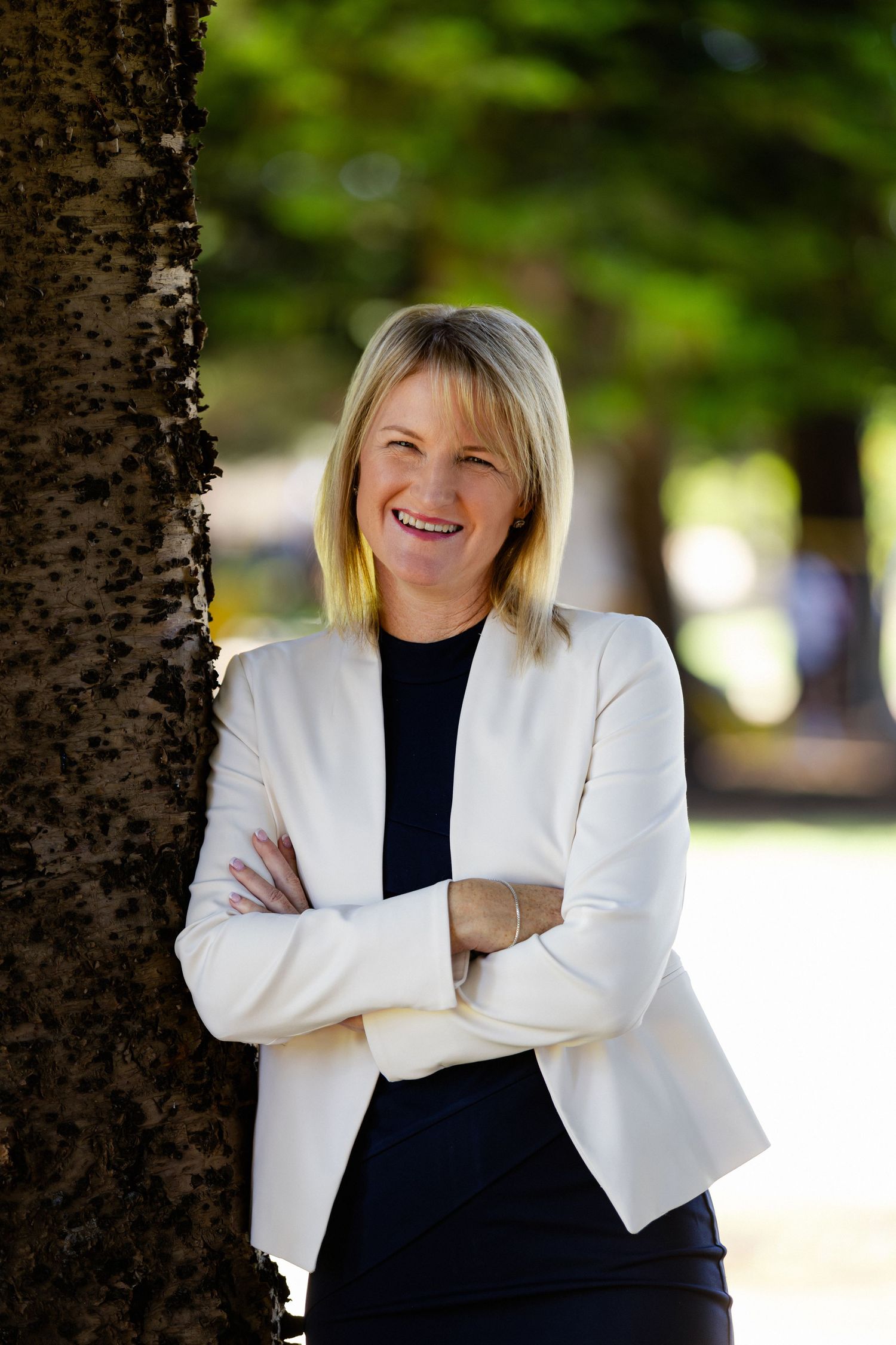 Professional portrait of someone wearing a white blazer and black top smiling against a blurred outdoor background.