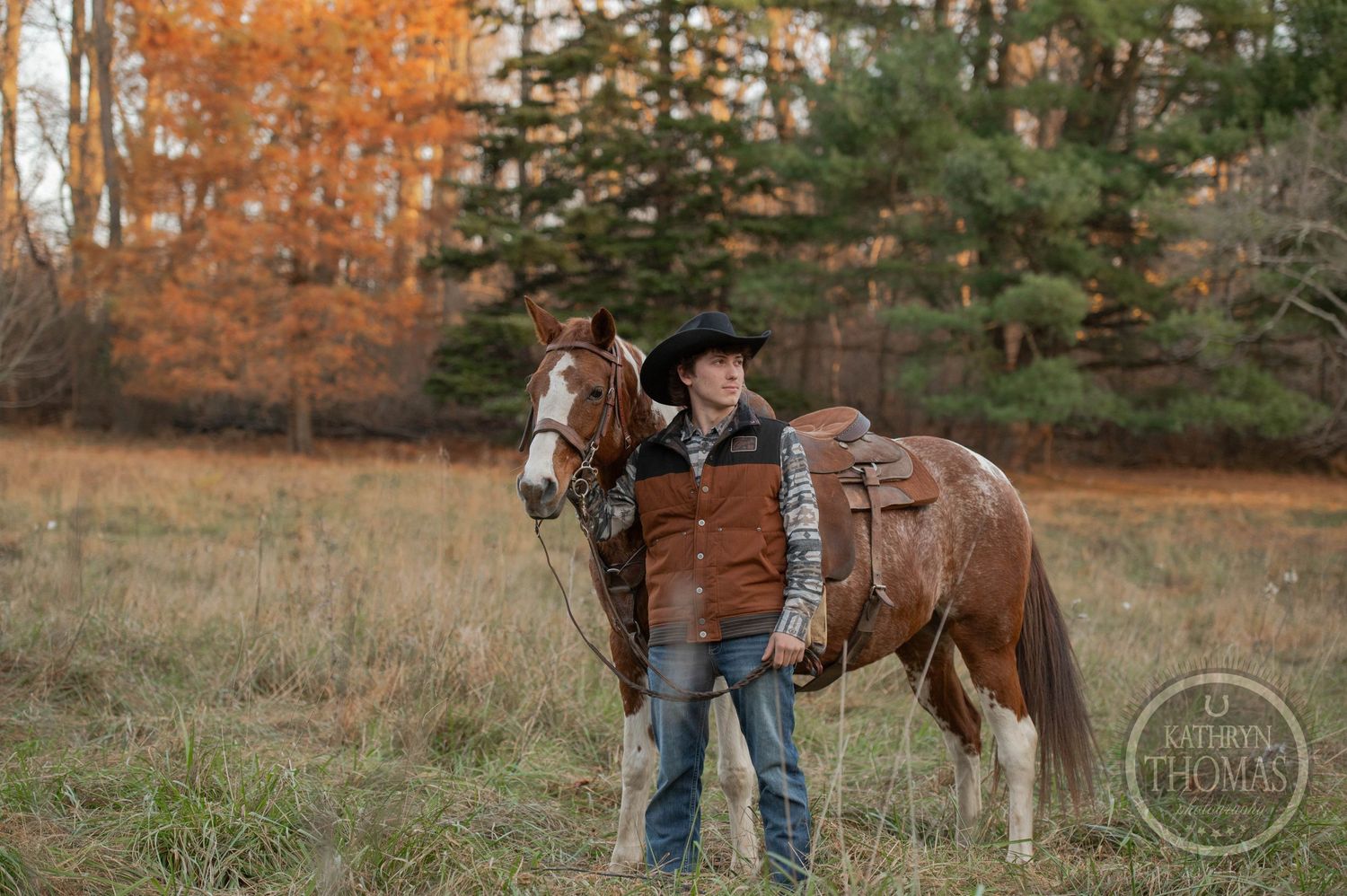 A paint horse with western tack stands next to a rider in brown vest and hat during fall season.