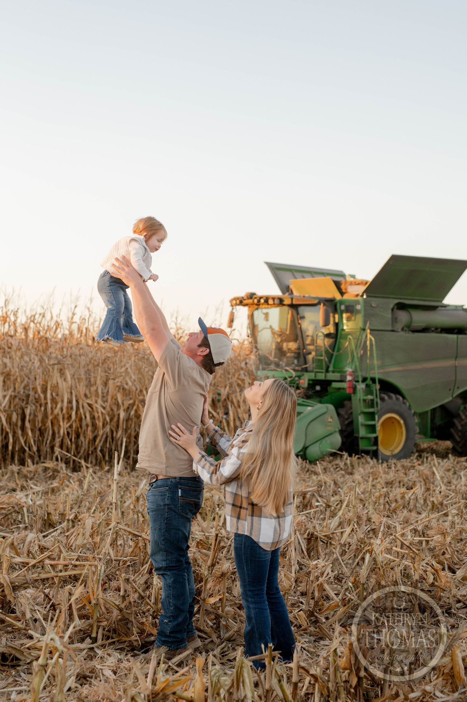 Parent lifts child in the air while playing in harvested cornfield with combine in background.