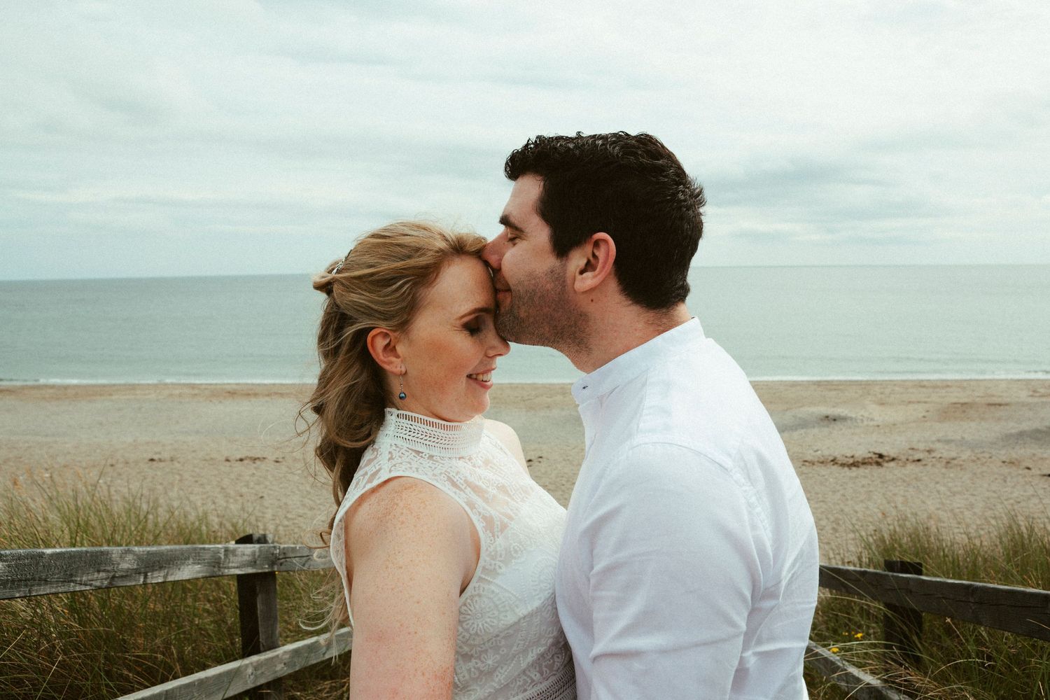 A romantic moment shared between a couple in white clothing on a coastal boardwalk overlooking the ocean.