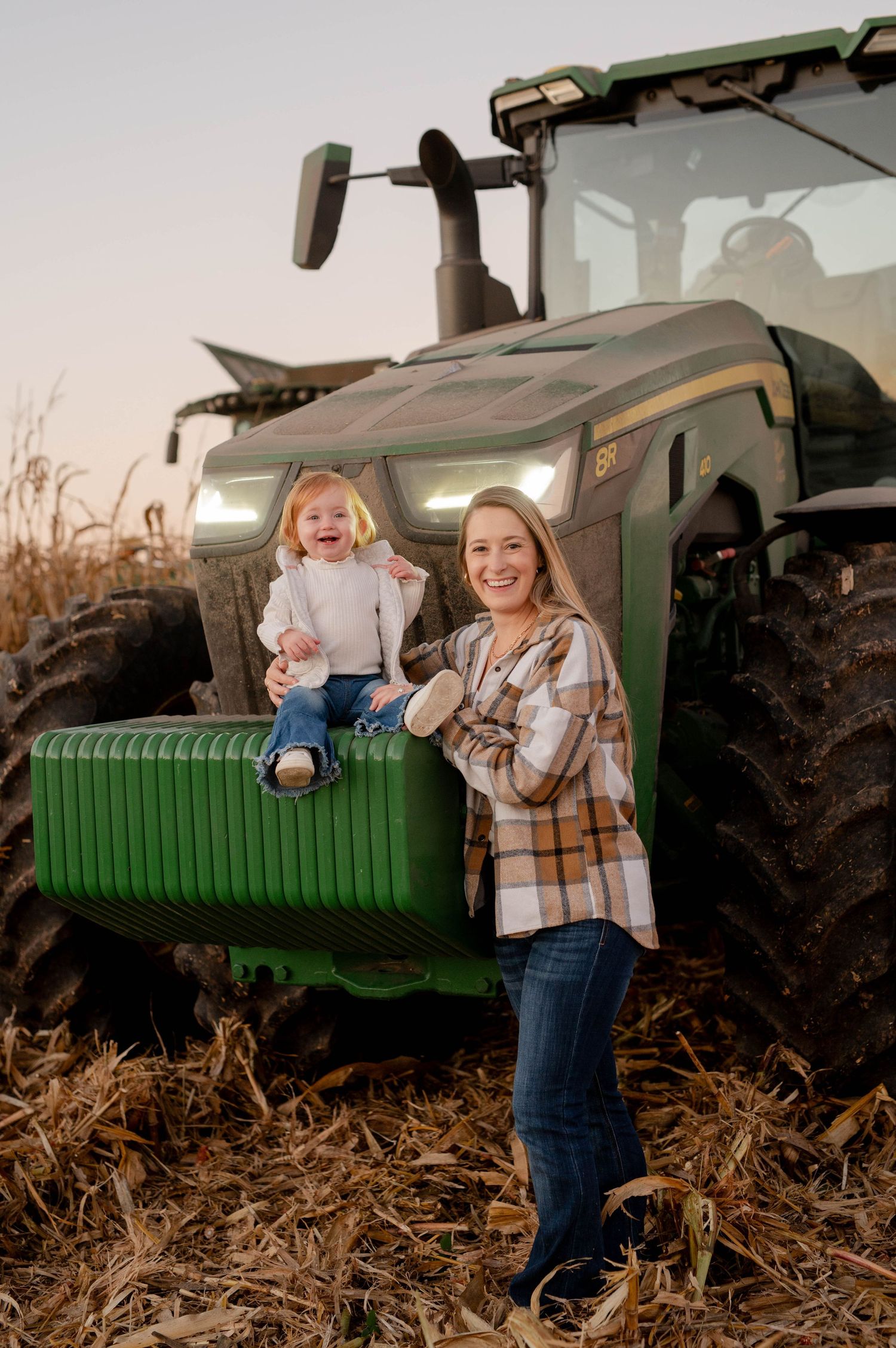 Mother and child pose together on a large green tractor in a harvested field.