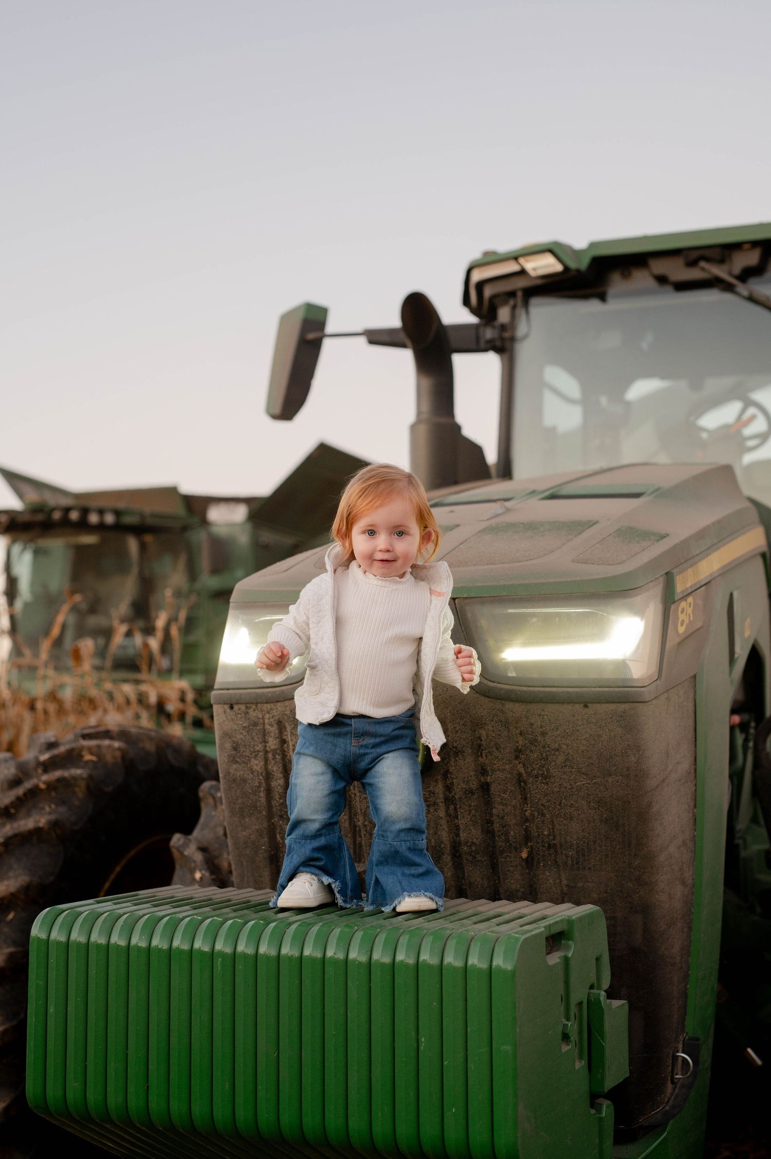 Young child standing on the front of a large green John Deere tractor at dusk.