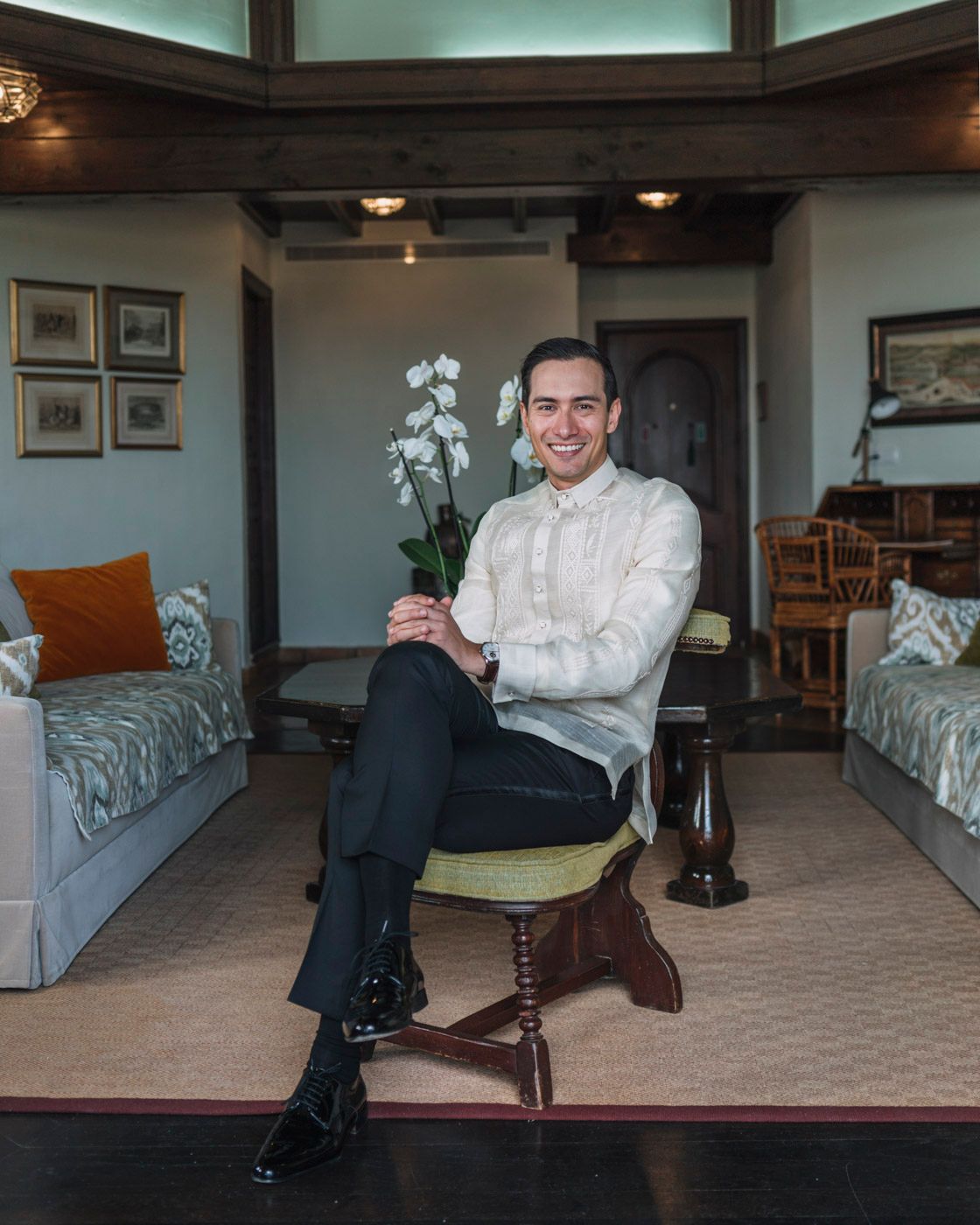 Person in a white dress shirt sits on a wooden stool holding a white orchid, with gray daybeds in the background.