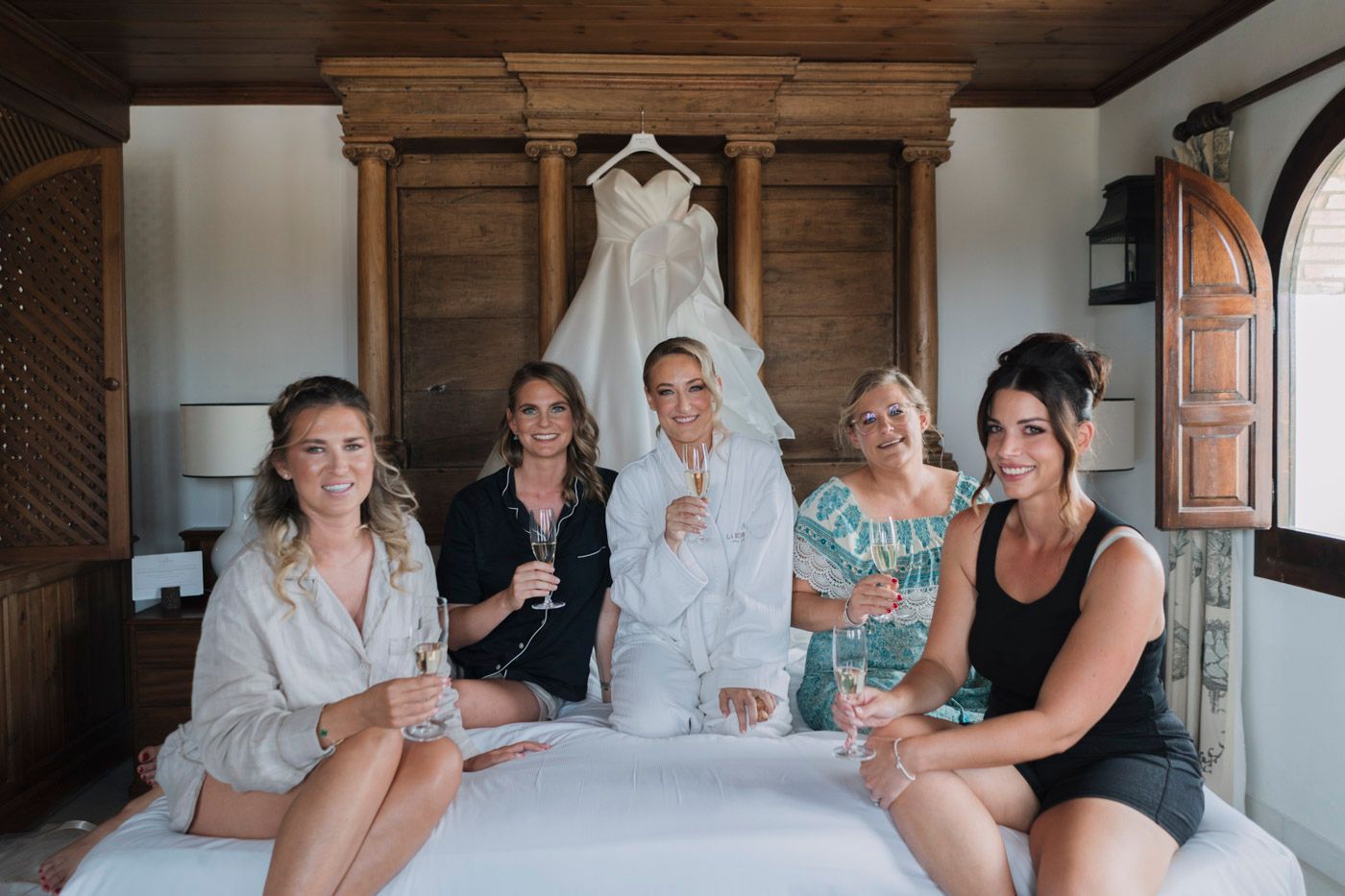 A white wedding dress hangs on display while a group relaxes together on a bed in a rustic wooden room.