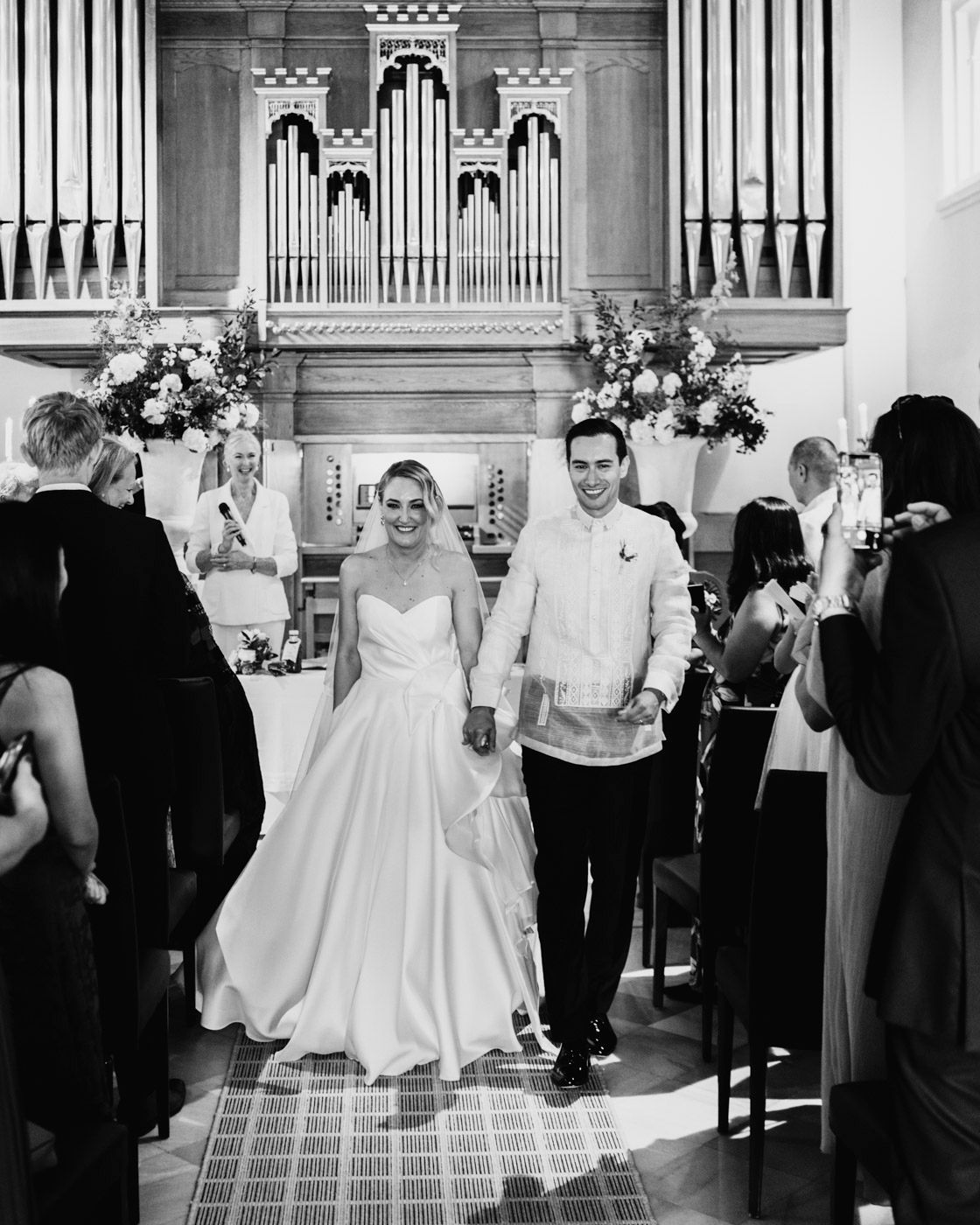 A couple walks down a church aisle as guests watch, with pipe organs and floral arrangements decorating the sanctuary.