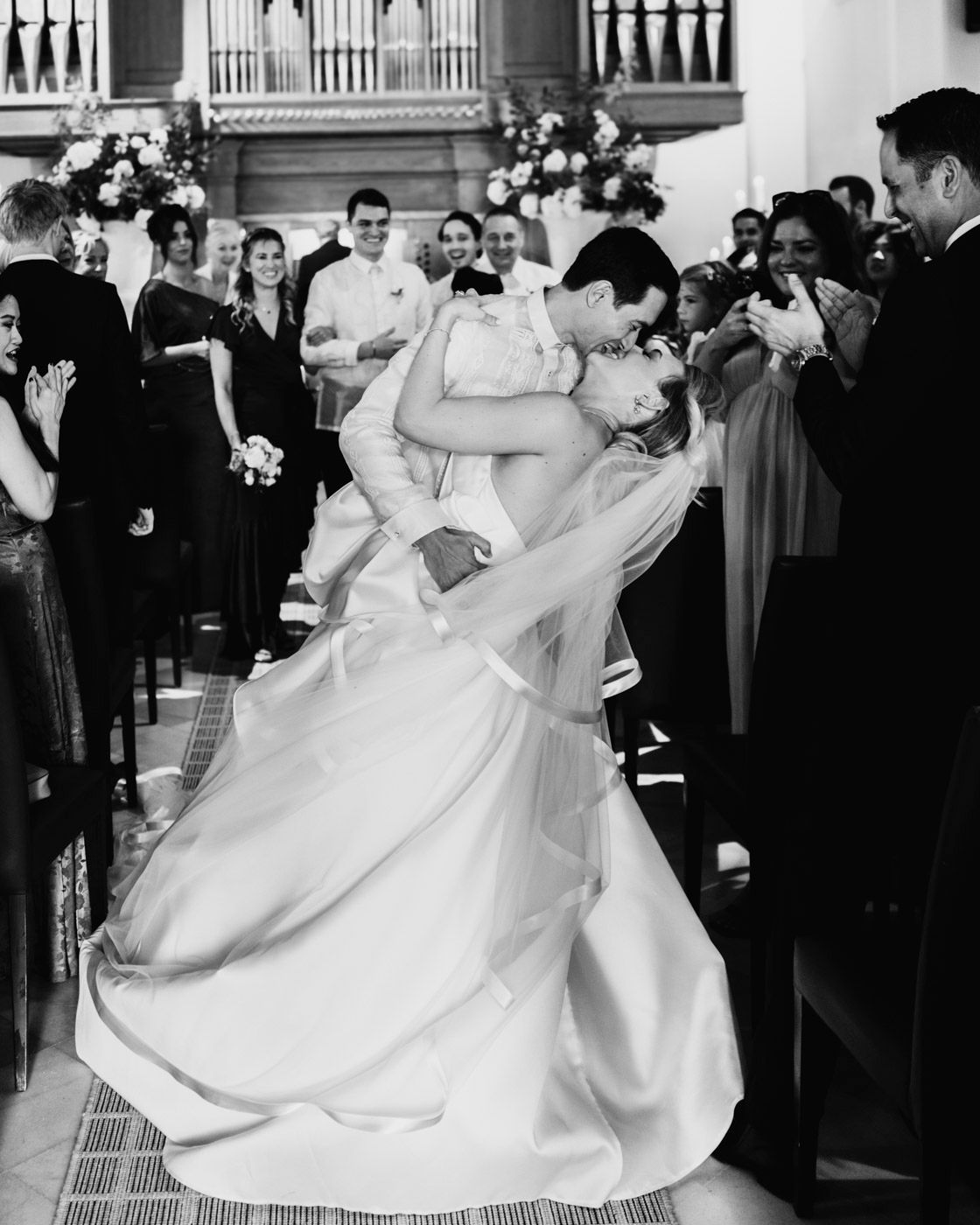 A romantic wedding couple shares a dramatic dip-style kiss while surrounded by guests in an elegant church setting.