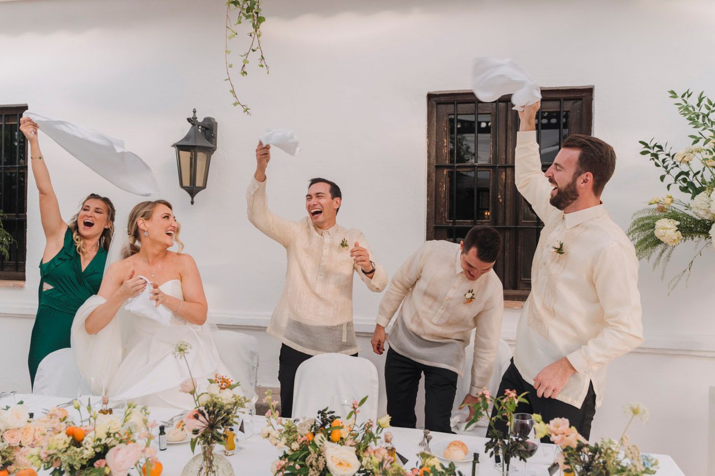Joyous wedding party celebrates with energetic dance moves at an outdoor reception table decorated with floral centerpieces.