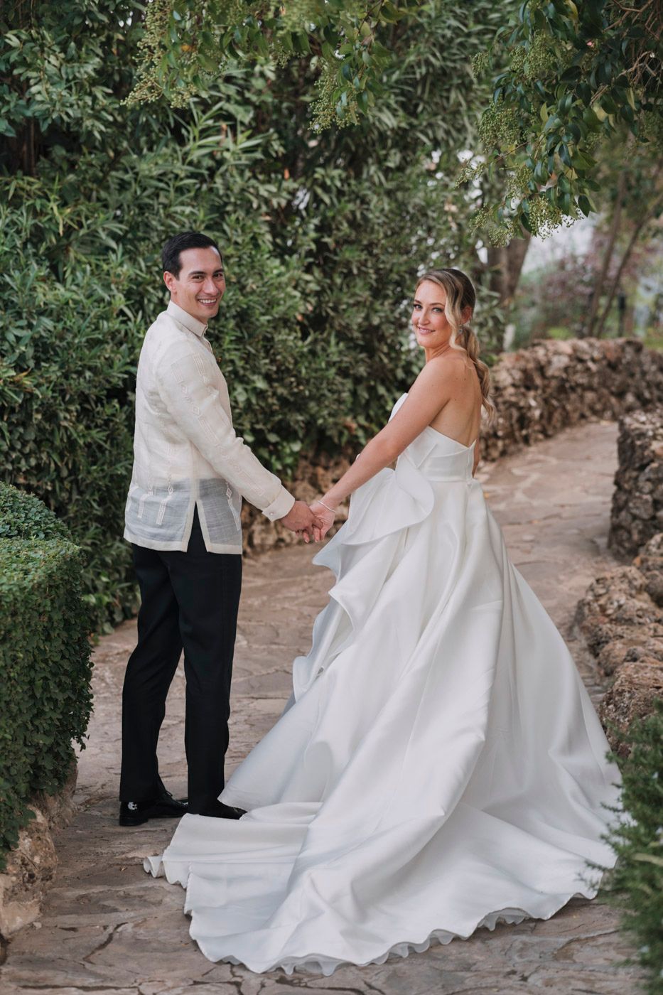 A couple in wedding attire pose on a stone path surrounded by lush greenery during their outdoor photoshoot.