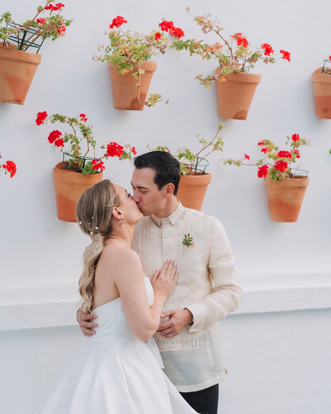 A romantic couple embraces against a white wall adorned with terracotta pots filled with blooming red geraniums.