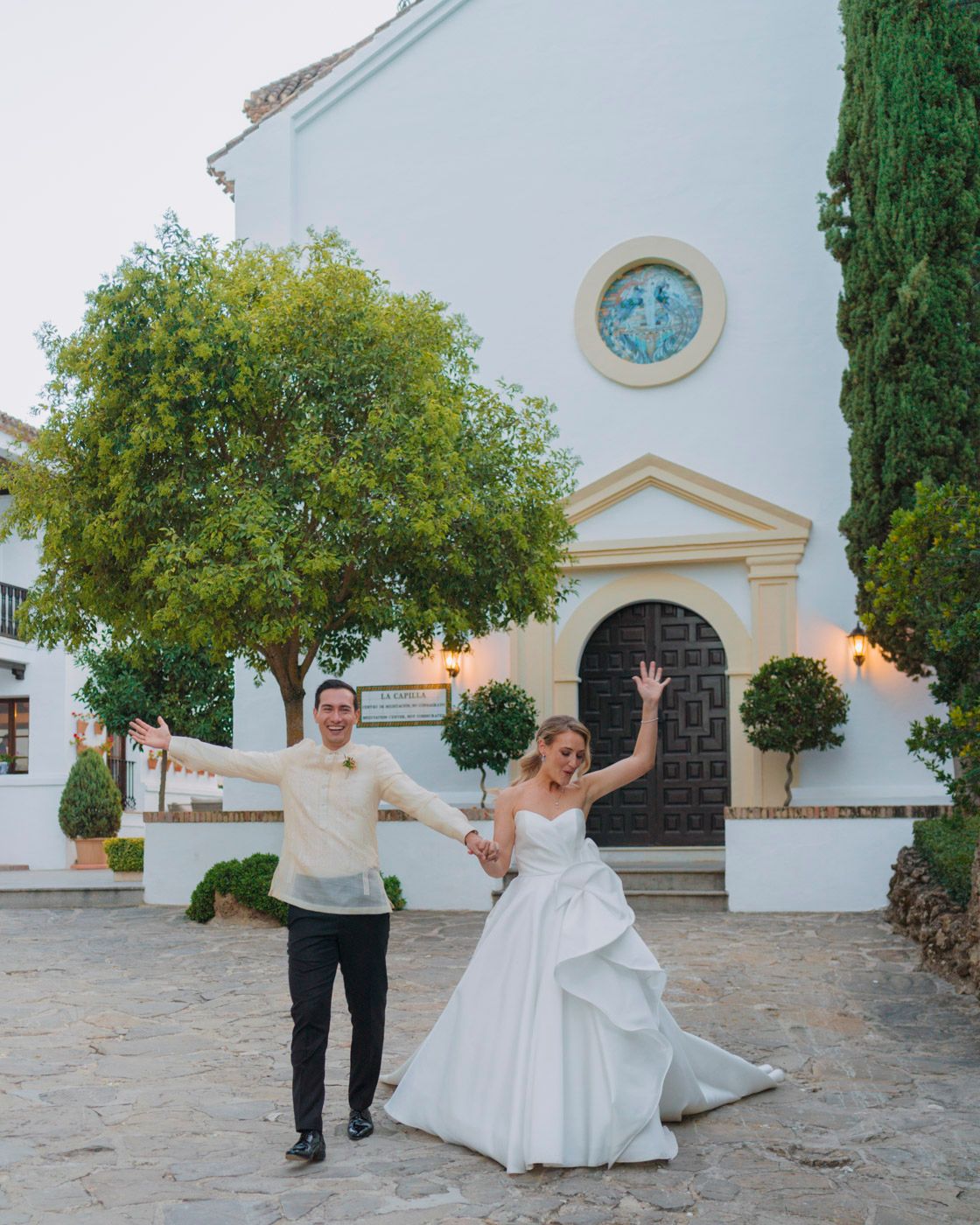 A joyful couple celebrating outside a white Mediterranean-style church with cypress trees and an arched entrance.