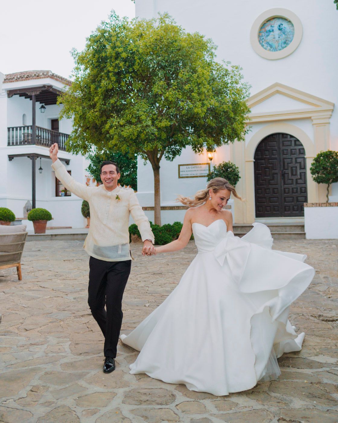 A joyful couple dances on stone tiles with a white Mediterranean-style building and courtyard in the background.