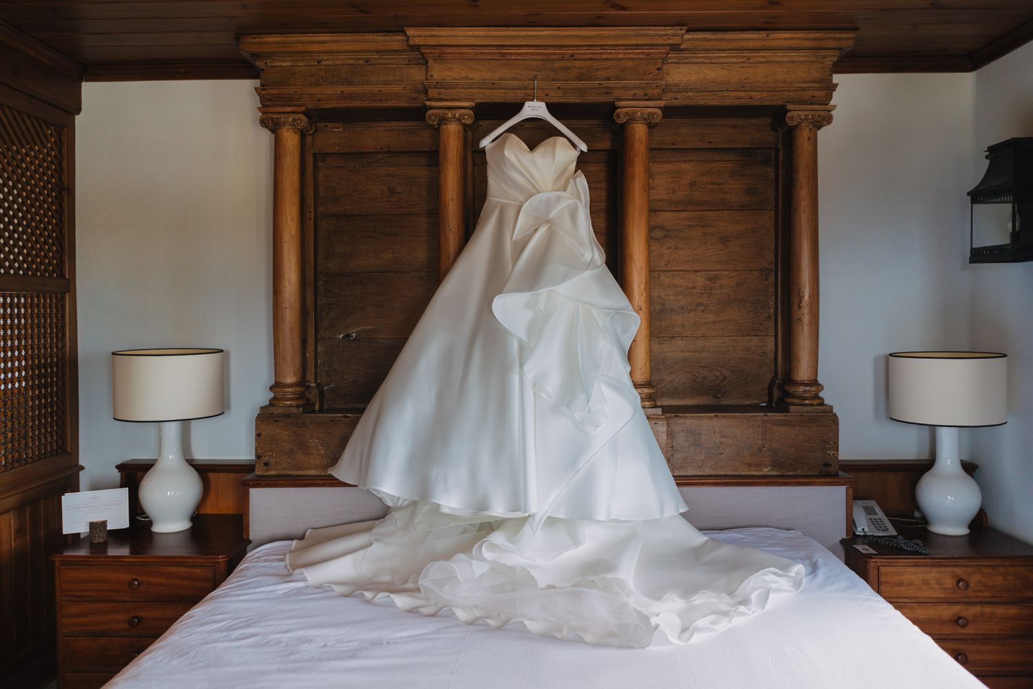 An elegant white wedding dress with flowing train hangs on display against a wooden wall between two matching table lamps.
