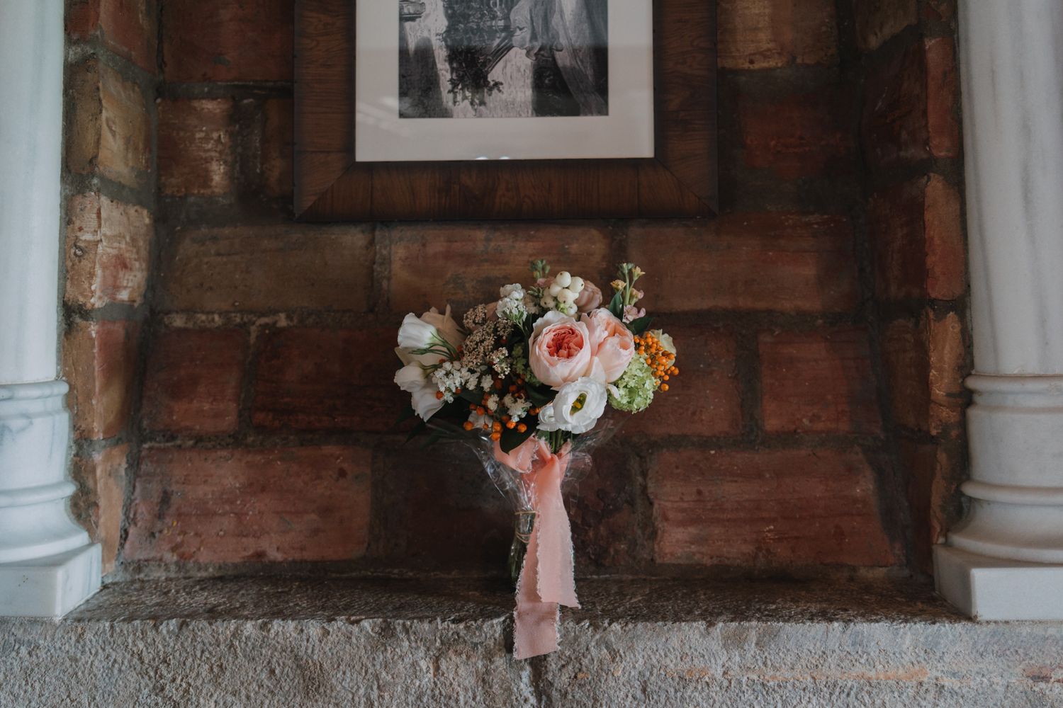A beautiful bridal bouquet with pink and white roses leans against a rustic brick wall beneath a framed photograph.