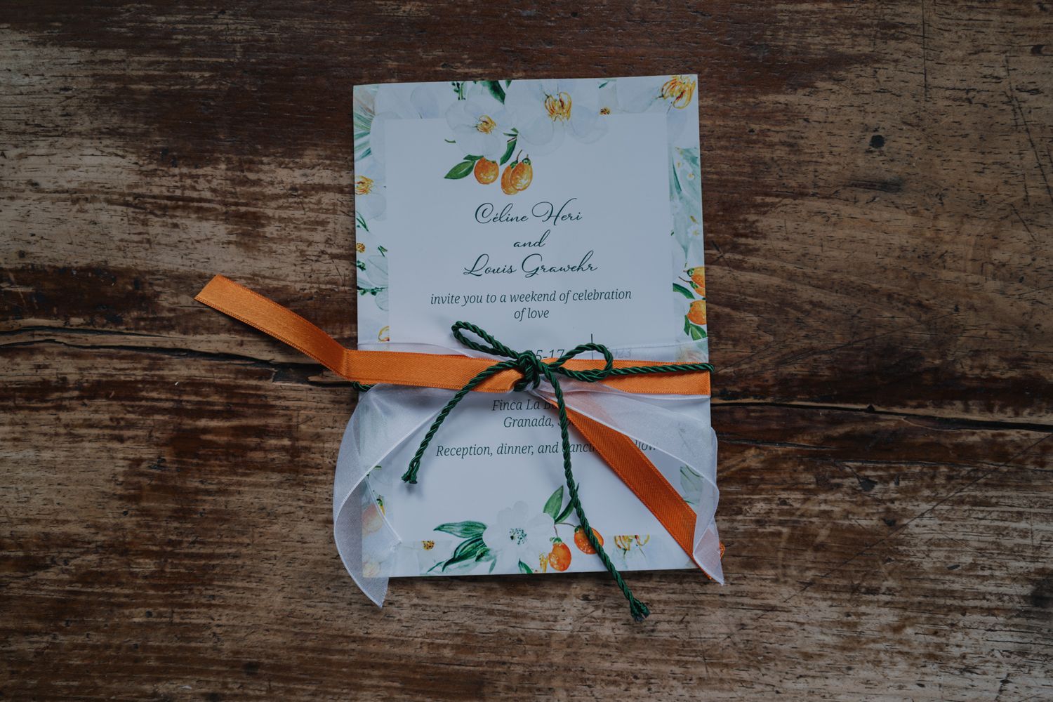 A floral wedding invitation tied with an orange ribbon bow sits on a rustic wooden surface.