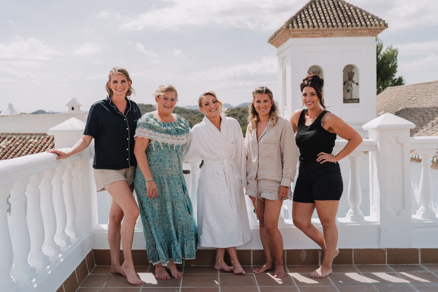 A cheerful group poses together on a Mediterranean-style balcony with white railings and a bell tower in the background.