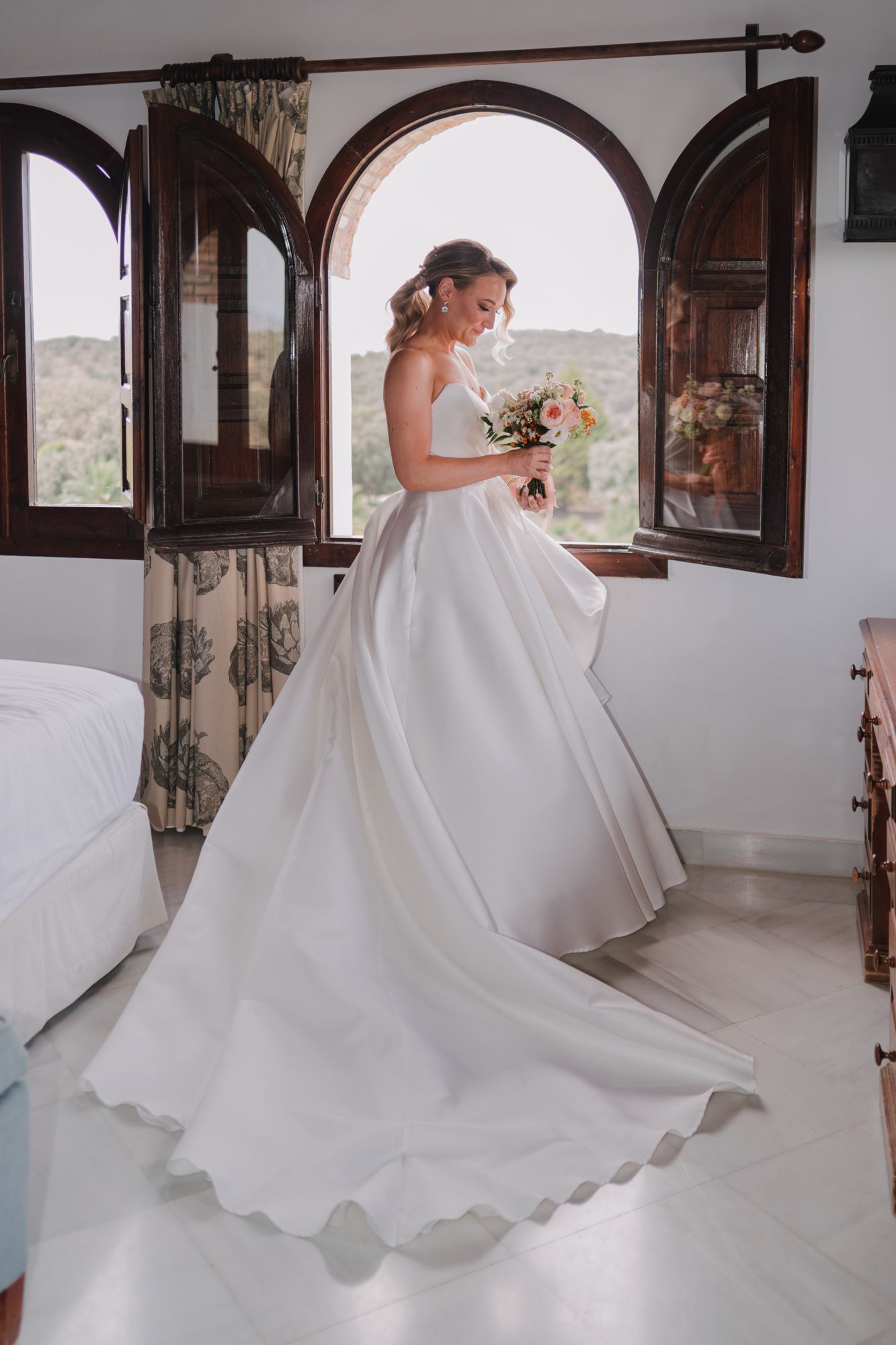 A bride in an elegant white ball gown with a flowing train stands by arched windows in a sunlit Mediterranean room.