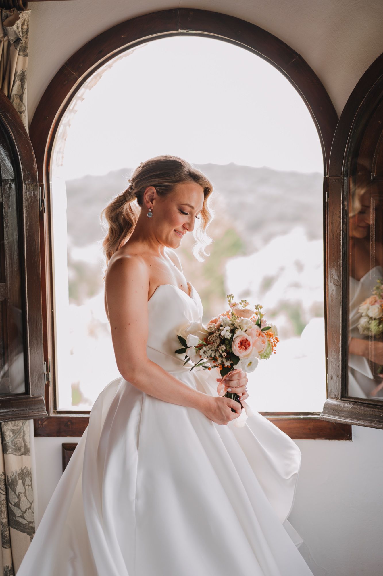 A bride in a classic white ballgown holds a pastel bouquet while standing in an arched window frame with natural light.