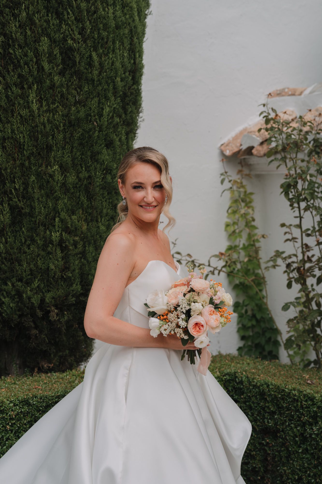 A bride in a white strapless wedding gown holds a peach and white floral bouquet against a garden backdrop.