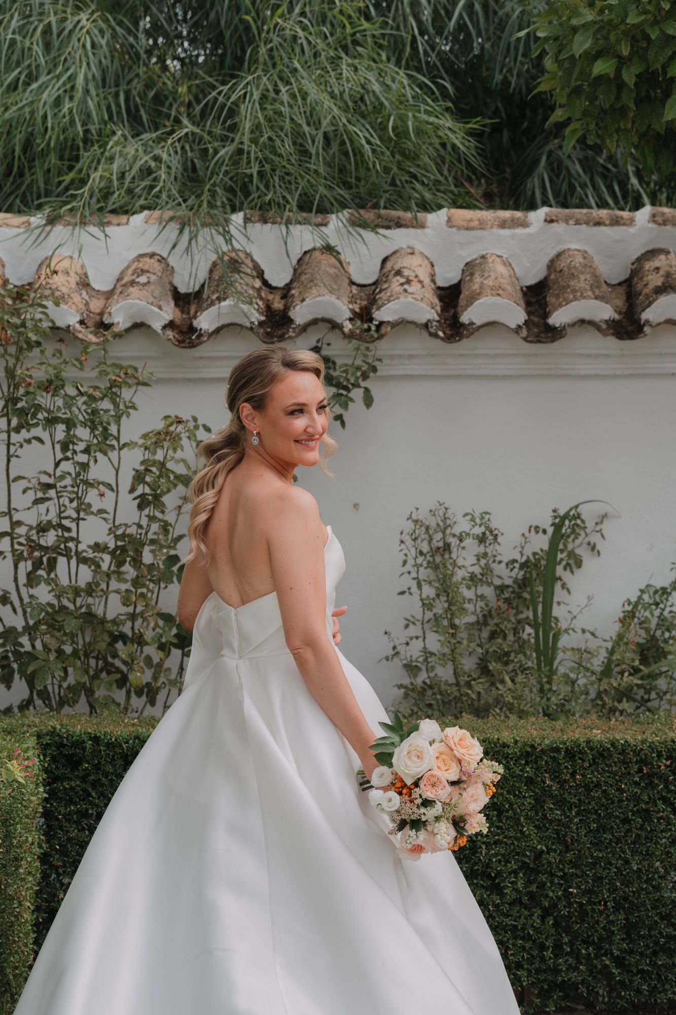 Elegant bride in strapless white ballgown holding peach-colored bouquet poses in garden with Spanish tile roof background.