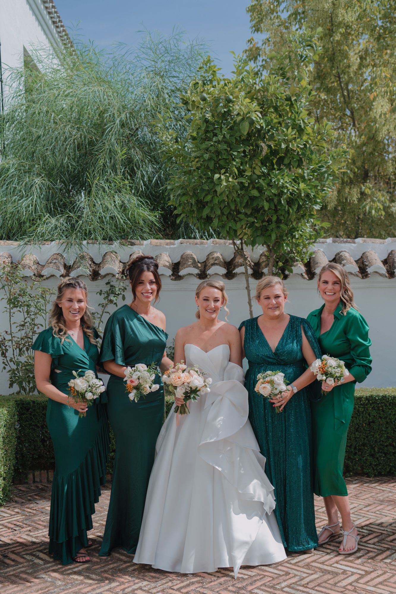 A bridal party poses together outdoors wearing coordinated emerald green dresses and holding white bouquets.