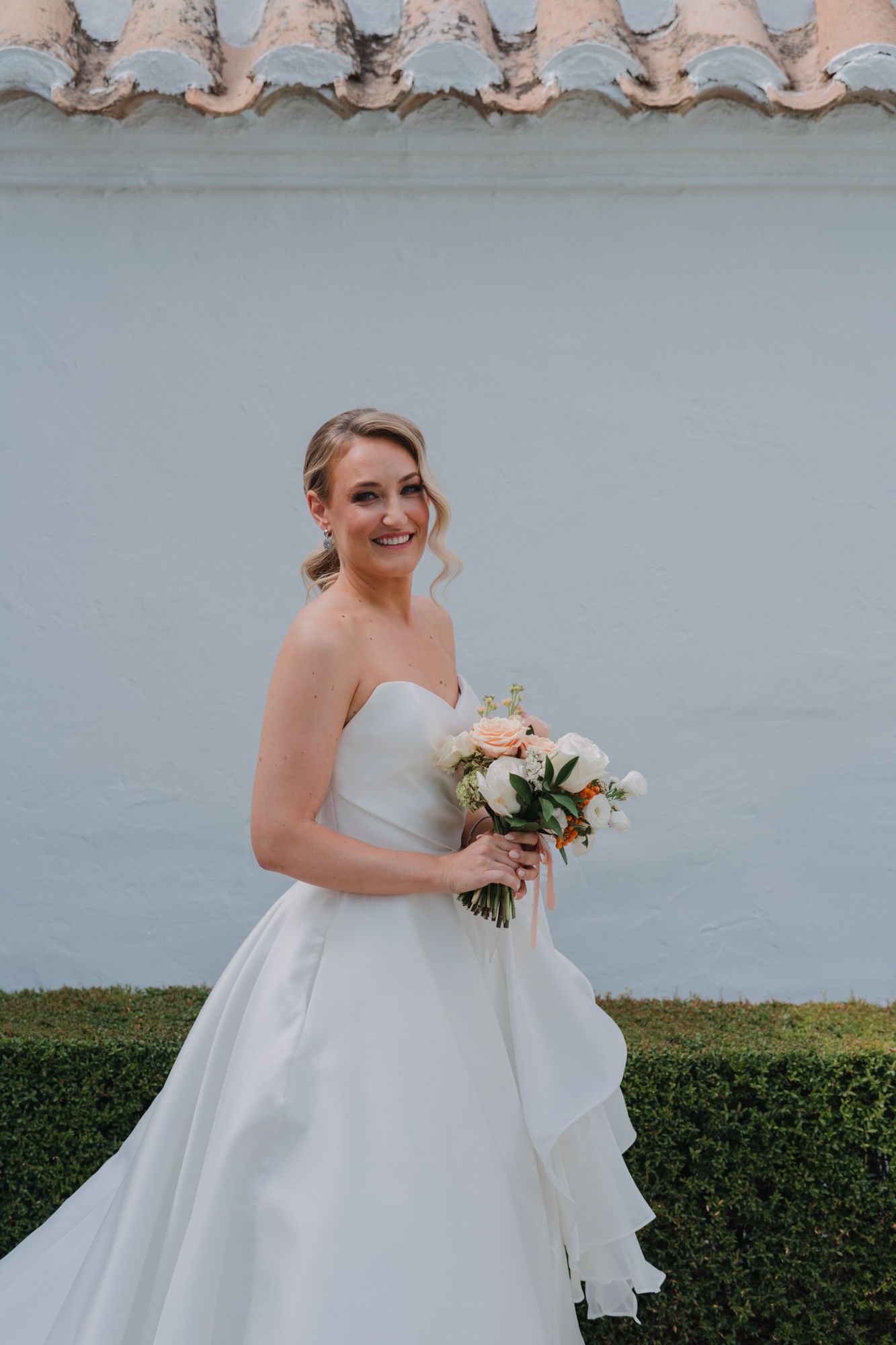A person in a white strapless ballgown wedding dress poses with a neutral floral bouquet against a gray wall.