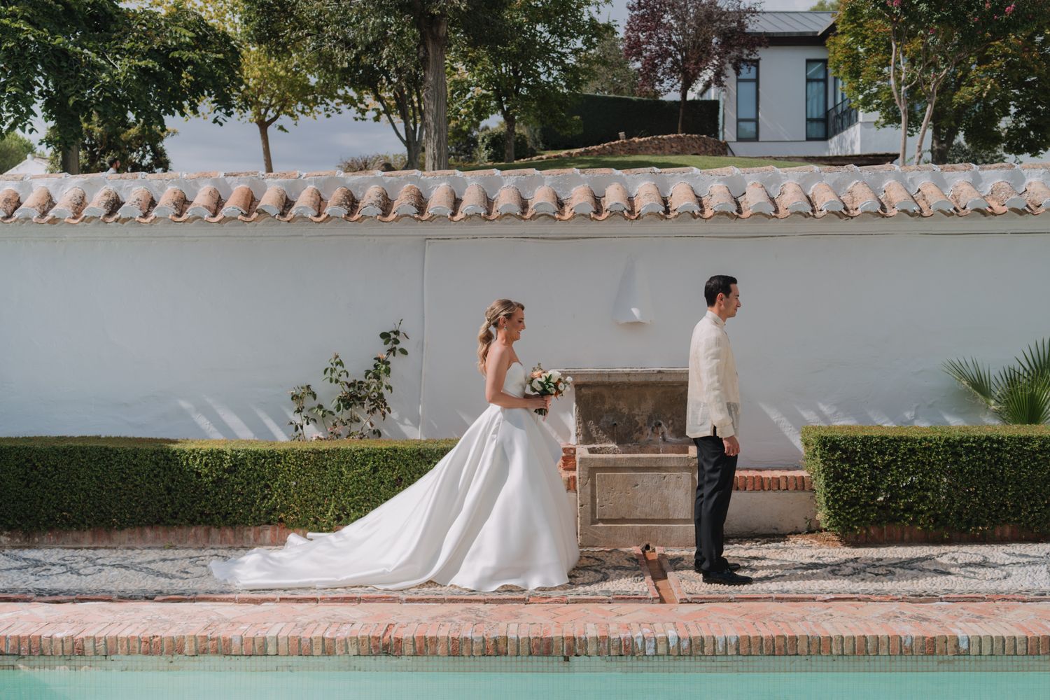 Elegant wedding couple poses beside white wall and manicured hedges near swimming pool.