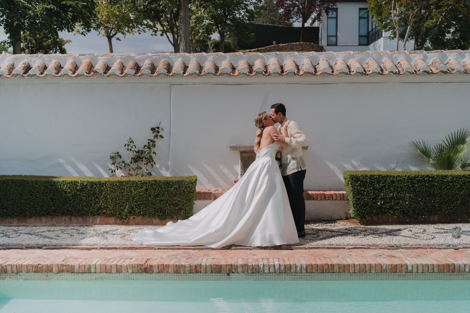 A romantic wedding portrait of a couple embracing by a swimming pool against a white stucco wall with terracotta tiles.