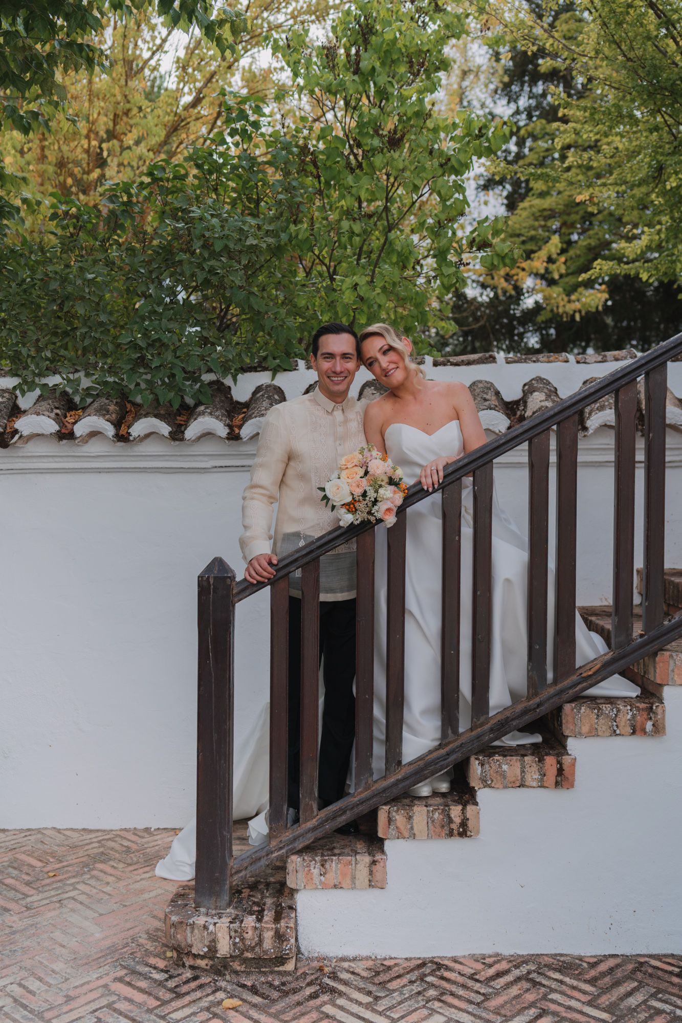 Wedding couple stands on stone staircase with white walls and lush greenery overhead.
