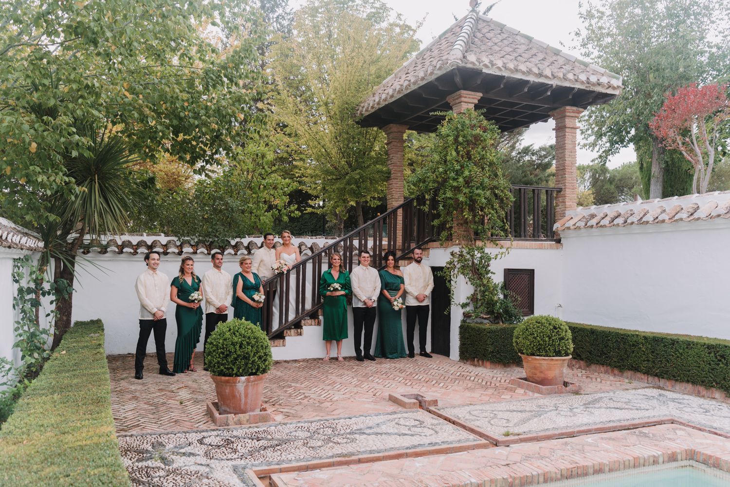 Wedding party gathered under wooden pergola in courtyard with white walls and landscaped gardens.
