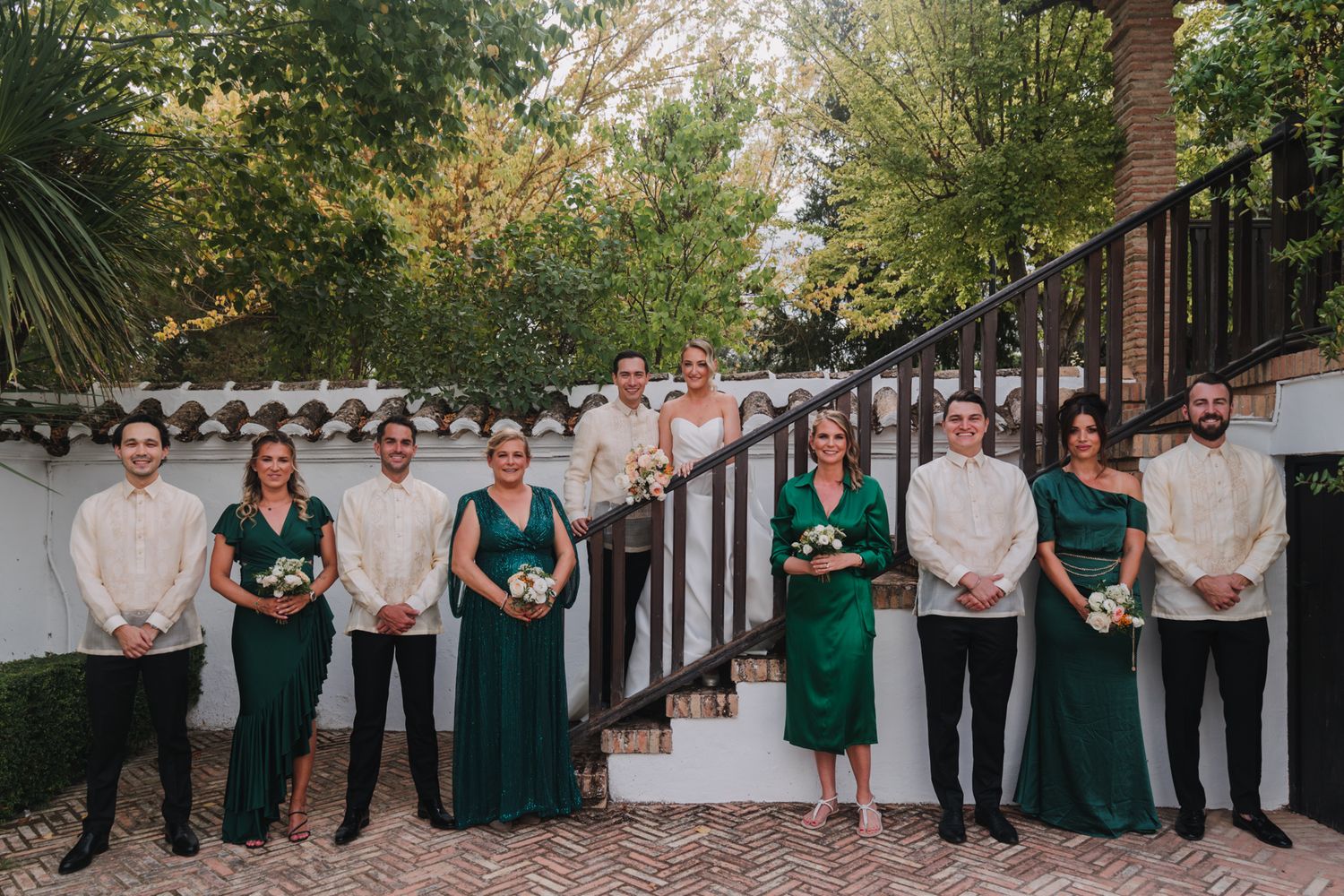 Wedding party in emerald green dresses and white shirts pose on outdoor stairs against white wall.