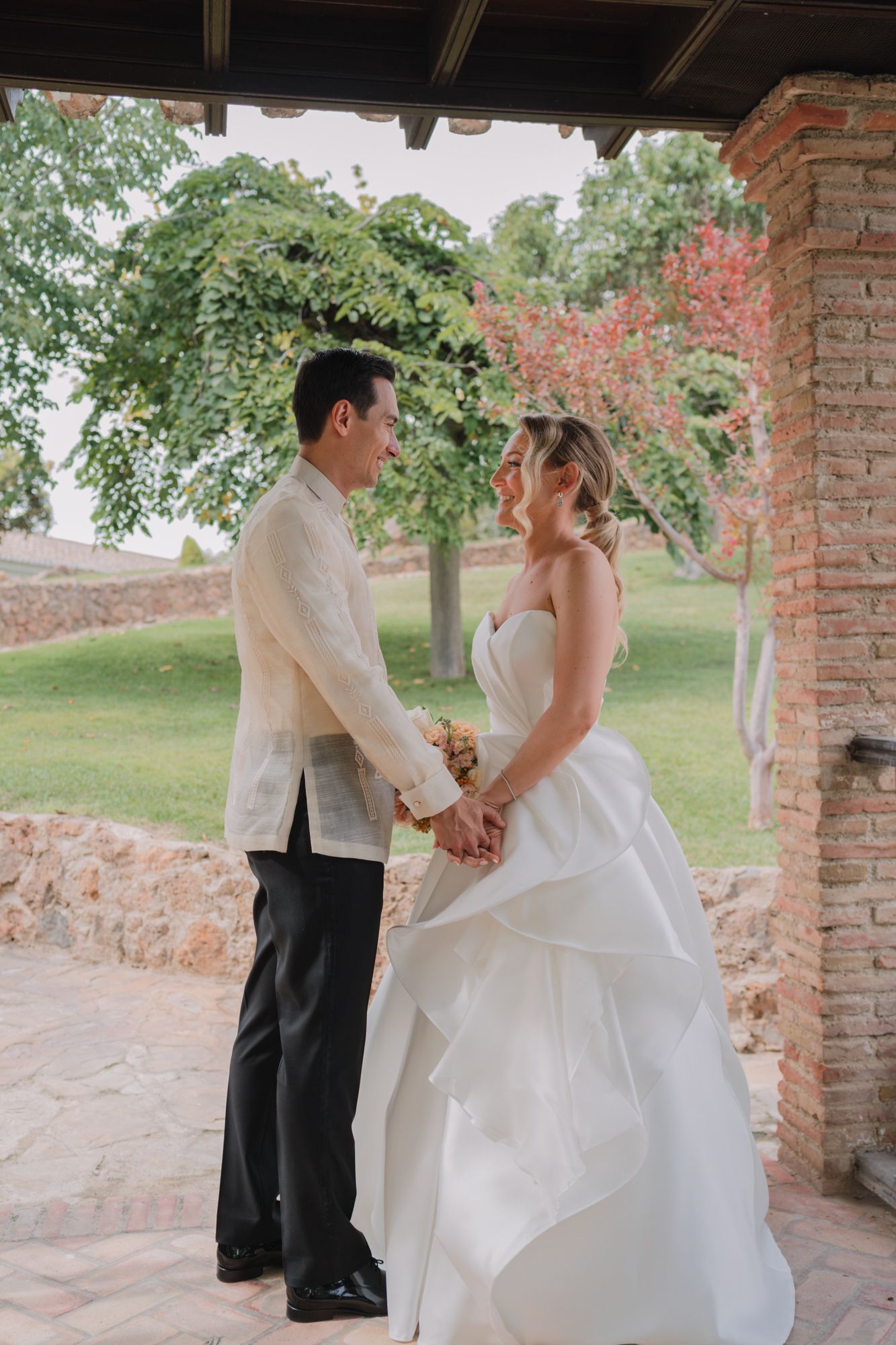 Wedding couple holds hands in a romantic garden setting framed by brick columns and colorful foliage.