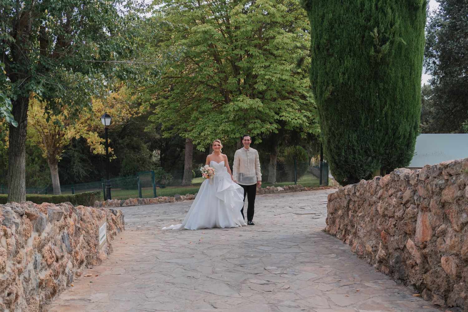 A couple walks along a stone-walled path surrounded by lush green trees and cypress in an elegant garden setting.