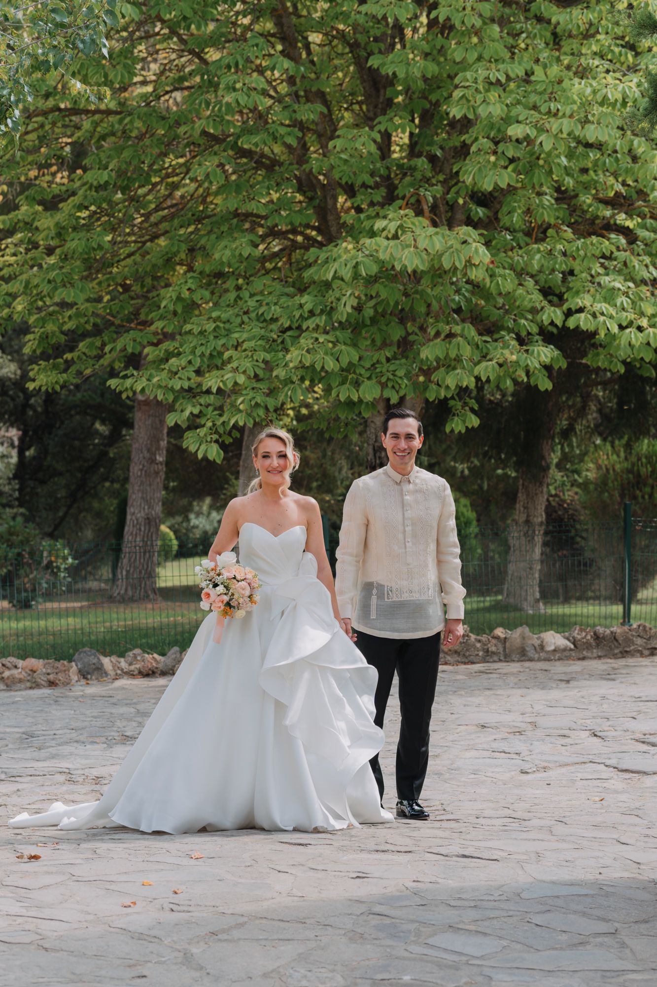 Wedding couple walks along stone pathway surrounded by lush green trees in an outdoor garden setting.