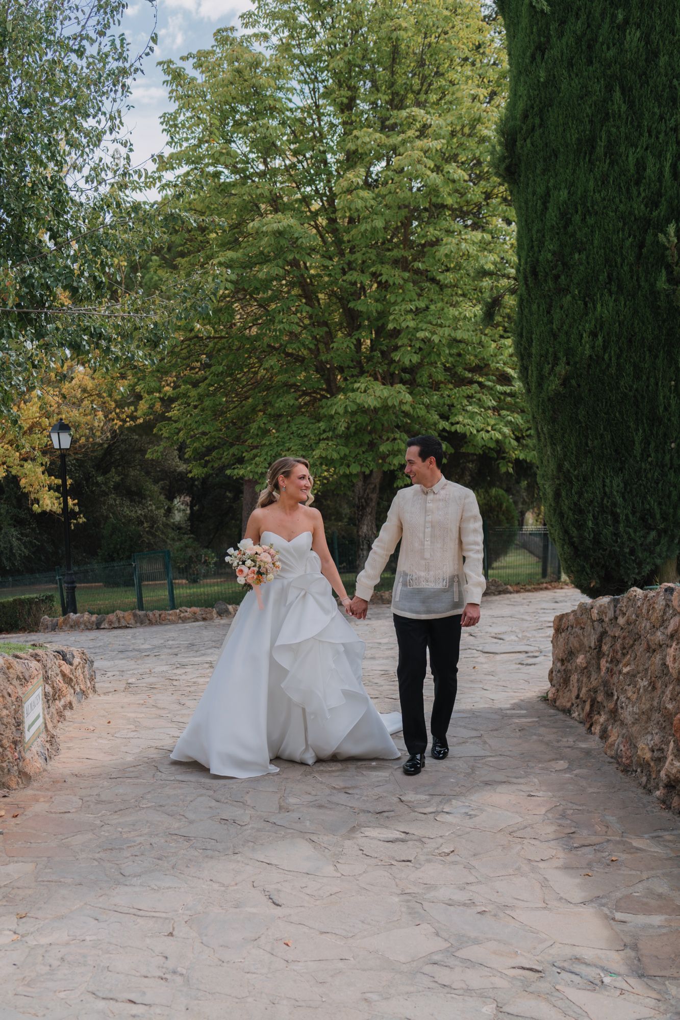 A wedding couple walks hand in hand along a stone path surrounded by lush green trees in a garden setting.
