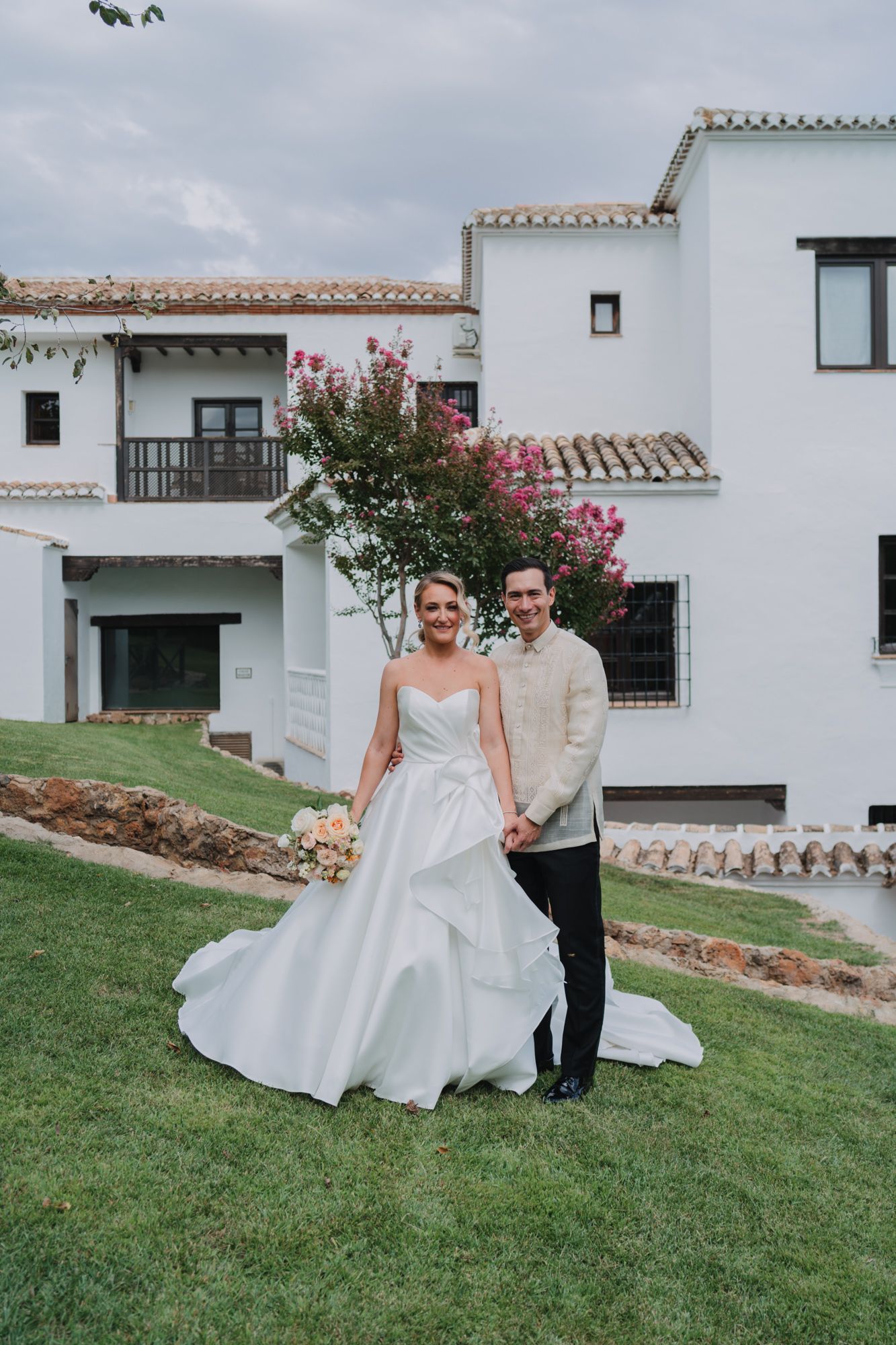 Two elegantly dressed wedding guests pose together on a grassy lawn in front of a white Spanish-style villa.