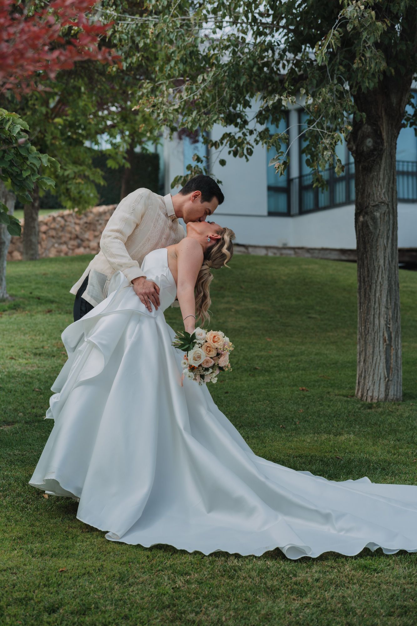 A romantic outdoor wedding photo of a couple dipping on a grassy lawn with a flowing white wedding dress and peach bouquet.