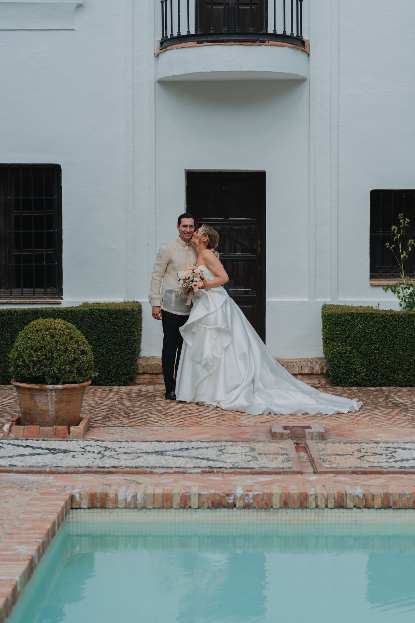 A bride and groom share an intimate moment by a poolside at an elegant white Spanish-style venue with manicured hedges.
