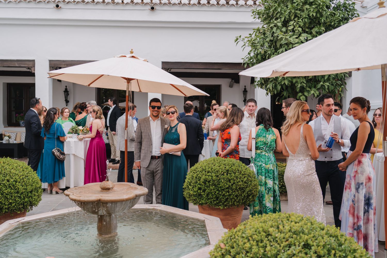 Wedding guests gather around an elegant stone fountain during an outdoor reception with cream umbrellas and manicured hedges.