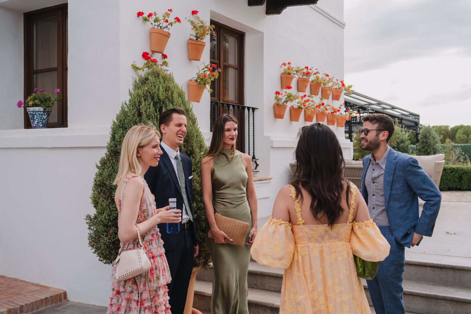 Group socializing at outdoor reception with white stucco walls and hanging flower decorations.